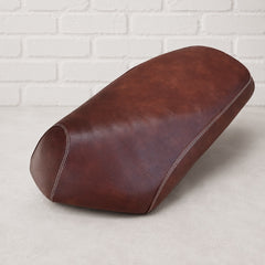 A brown leather scooter seat cover , shown from a three-quarters angle on a light gray floor against a white brick wall background. Made by Cheeky Seats for the Genuine Roughhouse in Pennsylvania